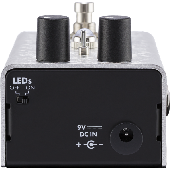 The image shows a compact guitar effects pedal with a silver metal housing, black control knobs, and professional input/output jacks. It features a 9V power input and LED indicators for status. Used on the floor and operated by foot, it modifies electric guitar sounds with effects like distortion or delay, serving as essential signal processing gear for live and studio use.