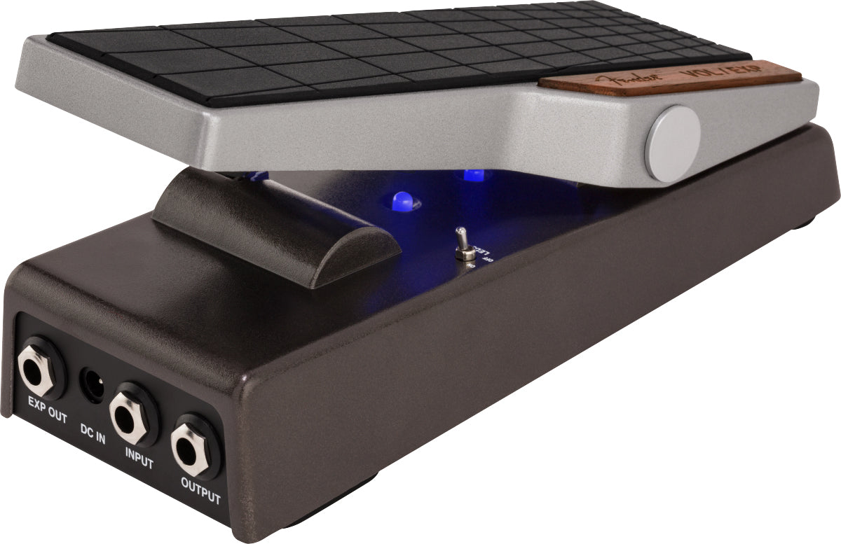 The image shows a modern wah pedal with a textured black rocker foot pedal, silver upper housing with a wooden accent, and a black lower housing featuring multiple 1/4" input/output jacks and a DC power input. Blue LED indicators light up when active. Wah pedals create a distinctive vocal-like "wah-wah" sound by sweeping frequencies using foot motion, widely used in rock, funk, and other genres since the 1960s.