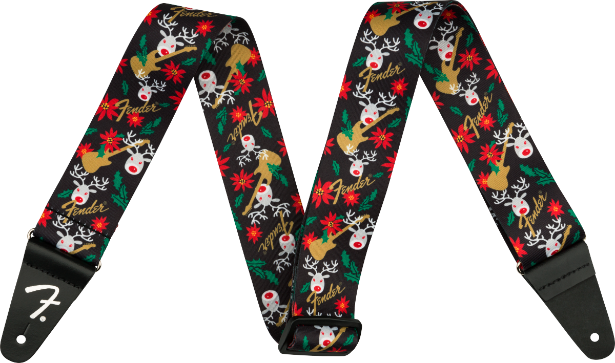 The image shows a Fender guitar strap with a black background featuring a festive pattern of red-nosed reindeer, red poinsettias, green holly leaves, white snowflakes, and gold Fender logos. It has black leather ends, one displaying the white Fender "F" logo, with holes for attaching to guitar strap pins. This seasonal accessory blends holiday designs with classic Fender branding.