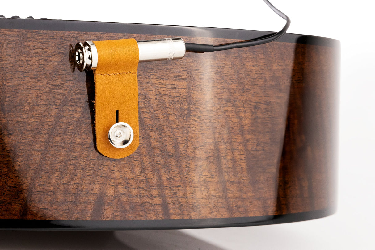 The image shows a contact microphone pickup mounted on an acoustic guitar’s body with an orange leather strap. The metallic cylindrical pickup has a cable for connecting to an amplifier or audio interface, allowing the guitar's sound to be amplified while preserving its natural tone. The guitar features a wooden body with visible grain, and the pickup appears professionally installed to capture vibrations effectively.