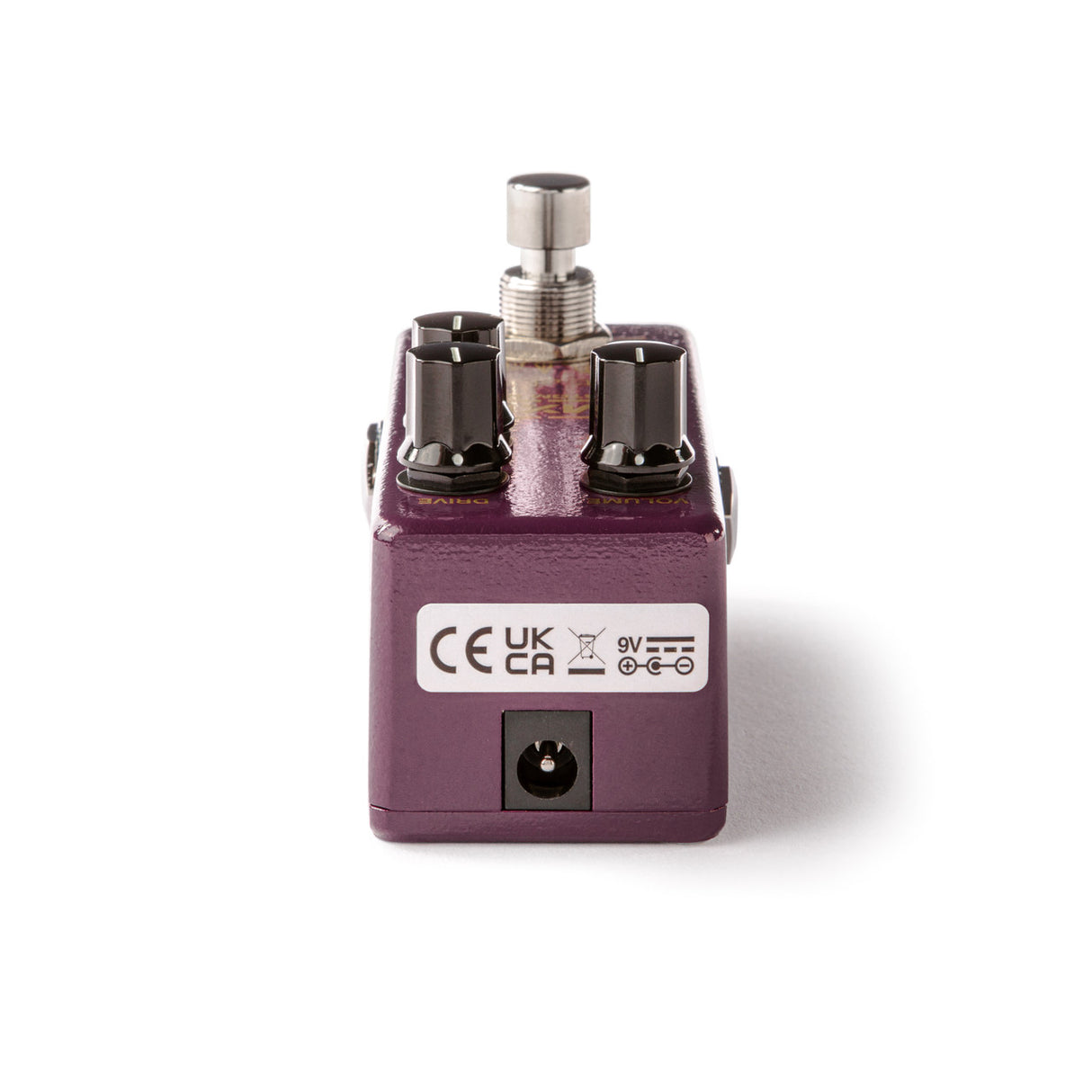 The image shows a compact guitar effects pedal with a purple/maroon metal housing. It features two input/output jacks, a control knob, a side power input for a 9V adapter, and CE certification marks. Designed as a stompbox, it modifies guitar sound with effects like distortion or delay and is built with sturdy metal typical of professional pedals.