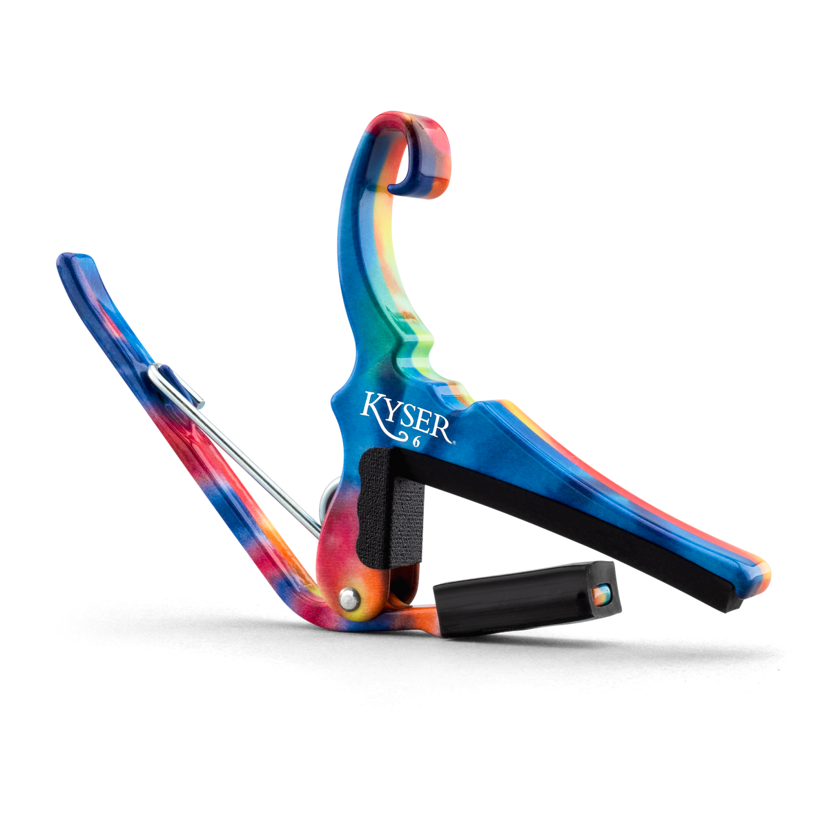 The image shows a colorful Kyser guitar capo with a vibrant tie-dye pattern featuring blues, reds, and oranges. It is spring-loaded with a curved clamp and has black padding to protect the guitar neck and strings. A small black cylindrical pick holder is integrated into the design. This accessory clamps the fretboard to change string pitch, combining functionality with a bright, artistic look.