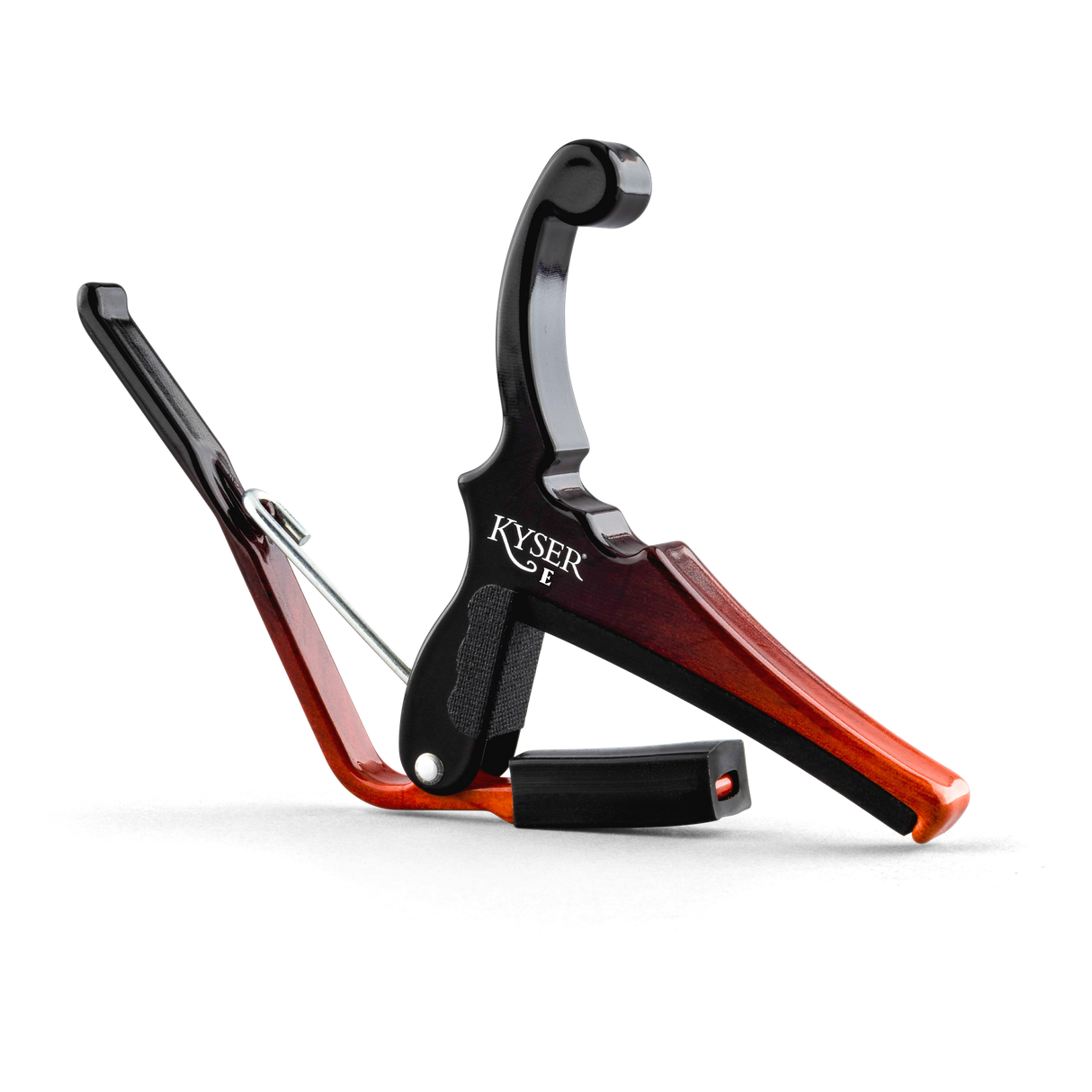 The image shows a Kyser guitar capo with a spring-loaded black metal clamp and a reddish-brown wooden finish. It features an ergonomic handle for one-handed use and black padded surfaces to protect the guitar neck and strings. Designed to clamp across the fretboard, it raises the pitch of all strings simultaneously, allowing quick key changes without retuning, making it popular for acoustic and electric guitars.