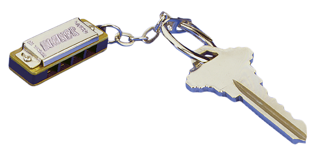 The image shows a miniature harmonica keychain attached to a key ring with a house key. The harmonica is small and functional, with a metallic silver finish and blue accents. It has a classic rectangular shape with holes on the front and the word "HARMONICA" embossed on the top. This novelty item serves as both a playable instrument and a keychain, popular as a compact musical accessory or gift.