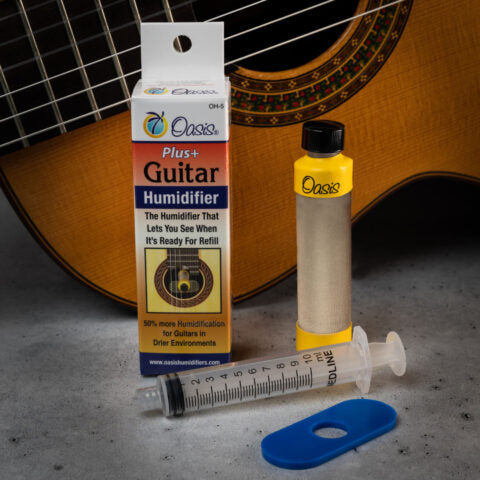 The image shows the Oasis OH-6 Plus+ Guitar Humidifier system placed in front of an acoustic guitar. The kit includes a cylindrical humidifier tube, a small bottle of yellow humidifying solution, a syringe for filling, a blue plastic clip, and the original packaging. The humidifier fits in the guitar’s sound hole to maintain proper humidity, protecting the wood from cracking and warping.