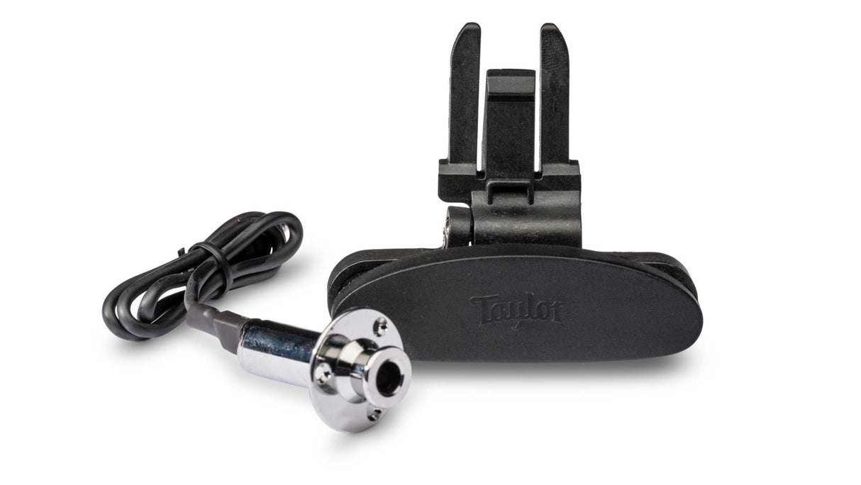 The image shows a Taylor acoustic guitar pickup and preamp system, featuring a black pickup unit, a branded control/preamp module with prongs, and a cable with a chrome 1/4" jack connector. This professional-grade system captures the guitar’s sound and allows amplification while preserving natural tone, commonly used by musicians for high-quality acoustic instrument amplification.