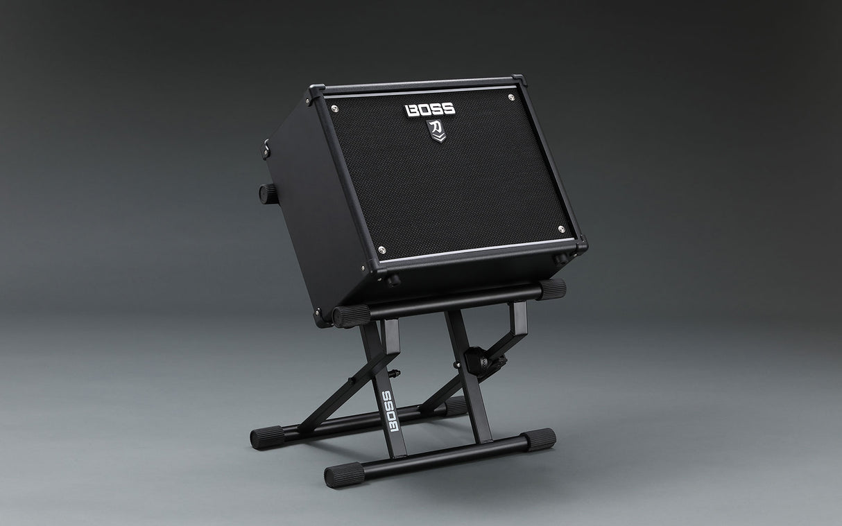 The image shows a compact black BOSS guitar amplifier mounted on an adjustable BOSS-branded stand. The amp features the distinctive BOSS logo on its front grille and is tilted for optimal sound direction. This portable setup is ideal for guitarists needing elevated amplification for better sound projection and easy control access, commonly used by both amateur and professional musicians.