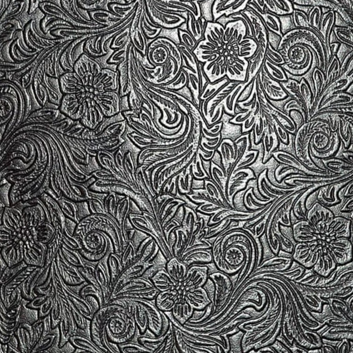 The image shows an ornate, tooled leather surface with intricate floral and scrollwork patterns in metallic silver against a dark background. This traditional embossed design is commonly found on musical instrument accessories like high-end guitar straps, vintage instrument cases, accordion bellows, banjo head surrounds, and premium music equipment bags, adding a functional yet elegant, vintage or western aesthetic.