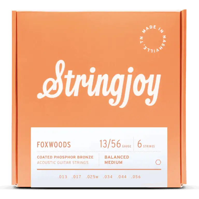 The image shows a package of Stringjoy Foxwoods coated phosphor bronze acoustic guitar strings. The orange/coral box features white lettering with the Stringjoy logo in script font. It’s a 6-string set, gauge 13/56, balanced medium tension, with gauges .013 to .056. A "Made in Nashville" badge indicates American-made quality. The coated strings offer warm tone and durability.