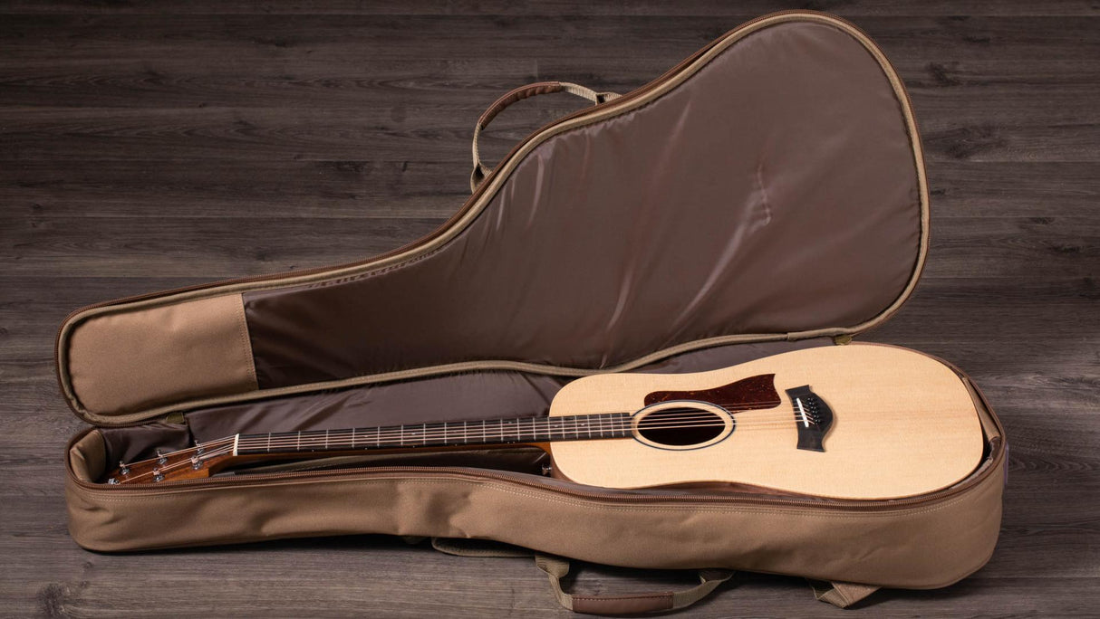 The image shows a steel-string acoustic guitar with a natural wood finish resting inside an open tan soft-shell gig bag. The guitar features a dreadnought-style body, a sound hole, and a dark pickguard. The padded case has zippers and handles and is placed on a dark wooden surface, suggesting a music-related setting like a studio or workshop.
