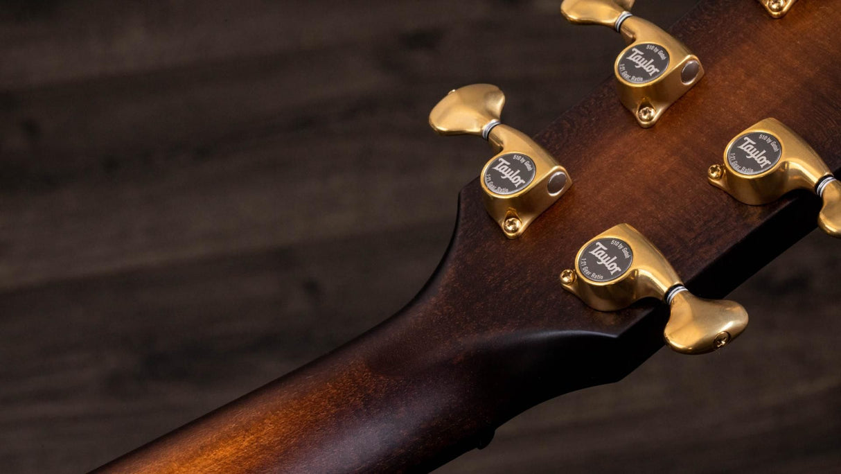 The image shows the headstock of a high-end acoustic guitar featuring four gold Taylor tuning machines. The rich brown wood grain, likely mahogany, highlights fine craftsmanship with smooth curves and professional finishing. The gold hardware combines functional precision tuning with elegant design, reflecting Taylor's reputation for premium, professional-grade acoustic guitars.