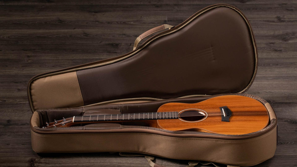 The image shows a steel-string acoustic guitar with a warm, natural wood finish resting in an open brown leather or leatherette hard case with plush interior padding. The case features a small storage compartment near the headstock. The guitar has a traditional dreadnought body shape and the setup is photographed against a dark wooden background, highlighting the instrument’s craftsmanship and protective case.