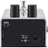 The image shows a compact guitar effects pedal with a silver metal housing, black control knobs, and professional input/output jacks. It features a 9V power input and LED indicators for status. Used on the floor and operated by foot, it modifies electric guitar sounds with effects like distortion or delay, serving as essential signal processing gear for live and studio use.