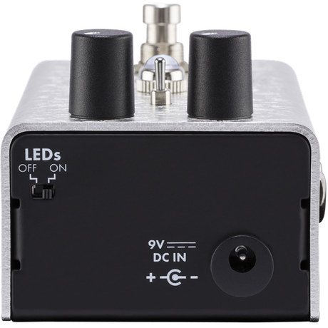 The image shows a compact guitar effects pedal with a silver metal housing, black control knobs, and professional input/output jacks. It features a 9V power input and LED indicators for status. Used on the floor and operated by foot, it modifies electric guitar sounds with effects like distortion or delay, serving as essential signal processing gear for live and studio use.