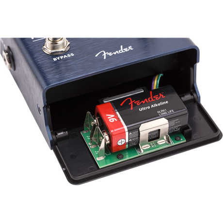 The image shows a Fender guitar effects pedal with a dark blue brushed metal finish and the Fender logo. Its battery compartment is open, revealing a green circuit board, a 9V Fender Ultra Alkaline battery with red and black labels, and connecting wires. Visible are the "BYPASS" switch and a 1/4" input jack. This typical setup illustrates the pedal’s internal electronics powering guitar effects either by battery or adapter.