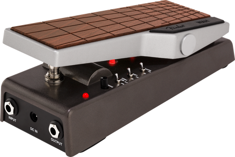 The image shows a guitar effects pedal, likely a wah pedal, with a hinged wood-grain treadle for foot control. It features toggle switches, a rotary knob, red LED indicators, 1/4" input/output jacks, and a DC power input. Housed in a durable metal enclosure, this pedal is used to create the classic "wah-wah" sound by filtering frequencies as the treadle rocks back and forth.