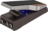 The image shows a modern wah pedal with a textured black rocker foot pedal, silver upper housing with a wooden accent, and a black lower housing featuring multiple 1/4" input/output jacks and a DC power input. Blue LED indicators light up when active. Wah pedals create a distinctive vocal-like "wah-wah" sound by sweeping frequencies using foot motion, widely used in rock, funk, and other genres since the 1960s.