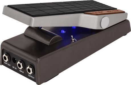 The image shows a modern wah pedal with a textured black rocker foot pedal, silver upper housing with a wooden accent, and a black lower housing featuring multiple 1/4" input/output jacks and a DC power input. Blue LED indicators light up when active. Wah pedals create a distinctive vocal-like "wah-wah" sound by sweeping frequencies using foot motion, widely used in rock, funk, and other genres since the 1960s.