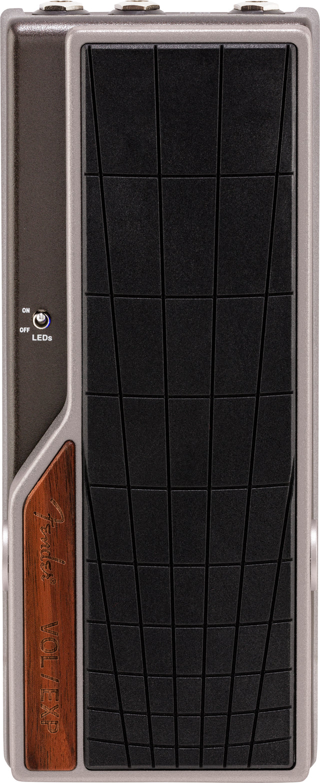 The image shows a Fender volume/expression pedal with a sleek gray metal housing and a large black textured footplate. It features the Fender logo on a wooden panel and an ON/OFF LED switch. The rocker-style design lets guitarists control volume or effects parameters like wah and delay for dynamic, expressive playing. Fender is a trusted brand known for quality guitar gear.
