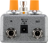 The image shows a guitar effects pedal with a gray textured metal enclosure. It features two orange-capped control knobs on top, labeled input and output 1/4" jacks on the front, a 9V DC power input, and an LED indicator light. This pedal modifies electric guitar sounds and is commonly used by guitarists to create effects like distortion, delay, and reverb in studio and live settings.