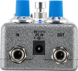 The image shows a gray, textured guitar effects pedal with two blue control knobs, a central metallic footswitch, and a small blue LED indicator. It has 1/4" input/output jacks on the sides and a 9V DC power input at the bottom. Designed for floor use, this compact pedal allows musicians to modify their instrument’s sound with various effects and is compatible with guitars, basses, and other electronic instruments.
