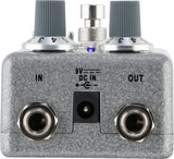 The image shows a metallic gray guitar effects pedal, likely a distortion or overdrive type. It features two control knobs, a central footswitch, and a small blue LED indicator. Input and output jacks are on the left and right, with a 9V DC power input in the center. The compact metal stompbox is designed for live use, altering guitar tone by adding distortion or overdrive in the signal chain.