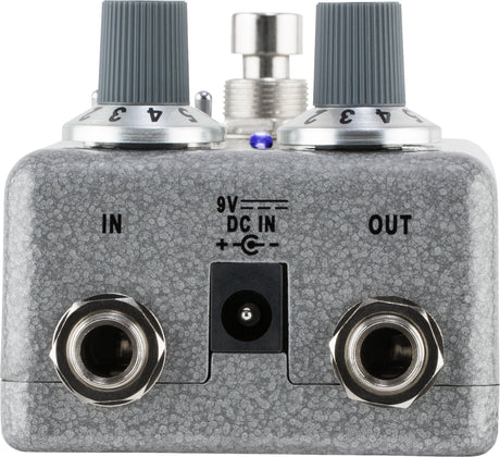 The image shows a metallic gray guitar effects pedal, likely a distortion or overdrive type. It features two control knobs, a central footswitch, and a small blue LED indicator. Input and output jacks are on the left and right, with a 9V DC power input in the center. The compact metal stompbox is designed for live use, altering guitar tone by adding distortion or overdrive in the signal chain.