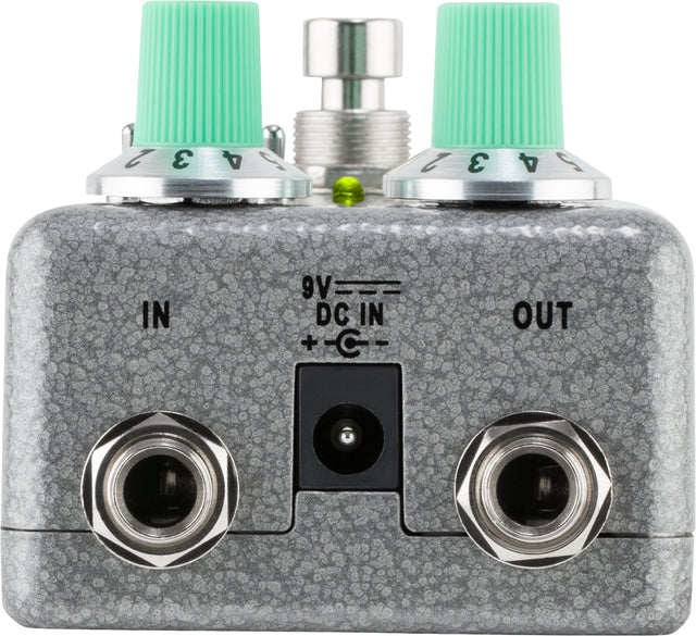 The image shows a guitar effects pedal with a compact, rectangular metal enclosure in speckled gray. It features two 1/4" jacks labeled "IN" and "OUT," a "9V DC IN" power input, two mint green control knobs, a chrome footswitch, and a small LED indicator. This pedal modifies guitar or bass sounds by processing the signal between the instrument and amplifier.