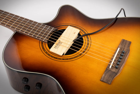 The image shows an acoustic-electric guitar with a sunburst finish, transitioning from dark edges to a golden center. It has a cutaway body, a wooden humbucker pickup branded "Teun Guitars" in the soundhole, a traditional rosette, steel strings, a rosewood fretboard, and a side-mounted control panel with knobs. An output cable is connected, highlighting its hybrid acoustic and electric functionality.