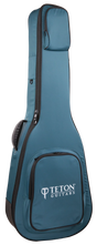 The image shows a teal/blue Teton Guitars padded gig bag shaped for acoustic guitars. It features the "TETON GUITARS" logo on the front pocket, multiple zippered compartments, padded shoulder straps, black zipper trim, and durable fabric. Designed for safe, lightweight transport, this soft case is ideal for carrying acoustic guitars and complements Teton’s branded instruments.