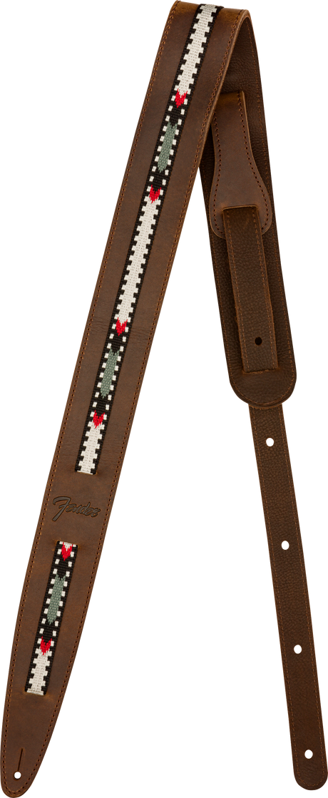 The image shows a brown leather guitar strap with visible stitching and a central woven fabric strip featuring a geometric pattern in white, black, green, and red. It has adjustment holes along its length and tapered ends for attachment. The "Fender" logo is visible, indicating the manufacturer. This strap combines functionality with a vintage, folk-inspired style.