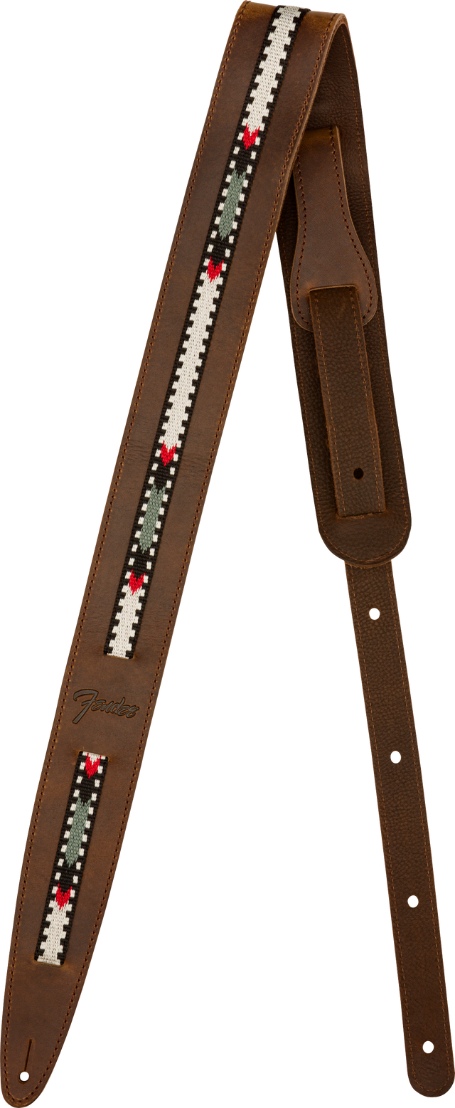 The image shows a brown leather guitar strap with visible stitching and a central woven fabric strip featuring a geometric pattern in white, black, green, and red. It has adjustment holes along its length and tapered ends for attachment. The "Fender" logo is visible, indicating the manufacturer. This strap combines functionality with a vintage, folk-inspired style.