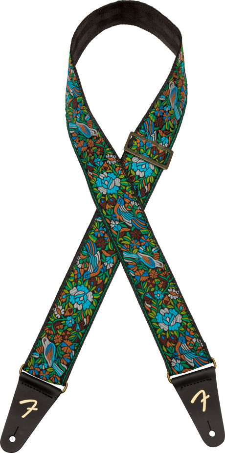 The image shows a vibrant Fender guitar strap with a detailed tropical pattern featuring birds, foliage, and bright colors like blues, greens, and browns. It has black leather ends embossed with the Fender "F" logo, metal hardware, and an adjustable buckle. The neck area includes black padding for comfort, combining both decorative style and functional support for playing guitar while standing.