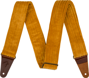 The image shows a guitar strap made of golden yellow corduroy with a ridged texture and brown leather ends, one embossed with a Fender logo. It features an adjustable metal slide buckle and a vintage-inspired design. This strap combines style and function, supporting the guitar when playing standing up while adding a retro, fashionable look.