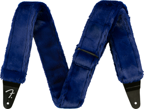 The image shows a guitar strap covered in luxurious blue fur or faux fur with black leather ends, one displaying the Fender logo. This plush, furry strap combines comfort and style, offering extra padding for long playing sessions while making a bold, eye-catching statement with its deep blue color and soft texture.