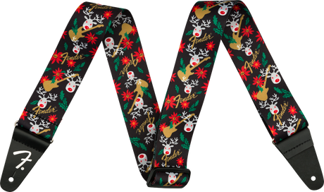 The image shows a Fender guitar strap with a black background featuring a festive pattern of red-nosed reindeer, red poinsettias, green holly leaves, white snowflakes, and gold Fender logos. It has black leather ends, one displaying the white Fender "F" logo, with holes for attaching to guitar strap pins. This seasonal accessory blends holiday designs with classic Fender branding.