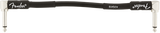 The image shows a black Fender guitar strap with the iconic Fender logo on the left side and additional branding on the right. It features white or silver metal hardware at both ends for attaching to a guitar’s strap buttons. This accessory helps musicians comfortably wear their guitar while standing by distributing the instrument’s weight across the shoulder and body.