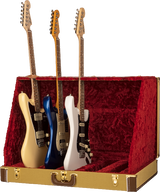 The image shows a vintage-style tweed guitar case or display stand with a red velvet interior holding three Stratocaster-style electric guitars. From left to right, the guitars are cream, blue, and white, each featuring single-coil pickups, tremolo bridges, maple necks with rosewood fretboards, and six strings. The case appears designed for elegant presentation or collector display.