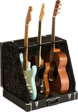The image shows a professional black hard case/display stand with plush lining designed to hold and display multiple guitars. It securely supports three guitars: a light blue Stratocaster-style electric, a black electric, and a natural wood dreadnought acoustic. The sturdy case features metal latches and neck supports, ideal for musicians, collectors, stores, or studios needing safe, accessible multi-guitar storage.