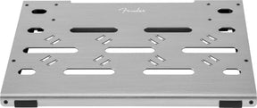 The image shows a Fender pedalboard, a flat, silver, brushed-metal platform with the Fender script logo. It features various slots and mounting holes for securing different guitar effects pedals, ventilation openings for heat dissipation, and rubber feet to prevent slipping. This sturdy, professional board helps guitarists organize and transport multiple pedals in a customizable, tidy setup.