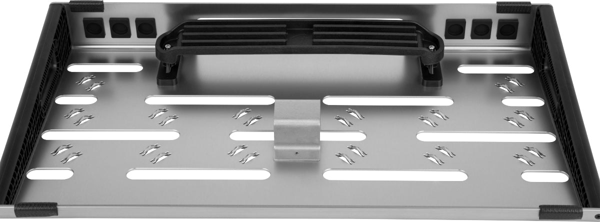 The image shows a rectangular guitar effects pedalboard with a metal surface, featuring white oval slots for securing pedals with velcro, a black carrying handle, protective black edges, and ventilation holes for airflow and cable management. Its sturdy construction is designed for easy transport and organizing multiple pedals in a customizable setup for performances or practice.