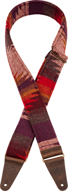 The image shows a guitar strap with a southwestern-inspired woven textile pattern in warm reds, burgundy, purple orange, and cream tones. It has brown leather end tabs with metal grommets for attachment and embossed branding. This decorative strap combines functionality—allowing the guitar to be worn standing—with a vintage, bohemian aesthetic that complements the player’s style.