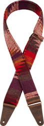 The image shows a guitar strap with a southwestern-inspired woven textile pattern in warm reds, burgundy, purple orange, and cream tones. It has brown leather end tabs with metal grommets for attachment and embossed branding. This decorative strap combines functionality—allowing the guitar to be worn standing—with a vintage, bohemian aesthetic that complements the player’s style.