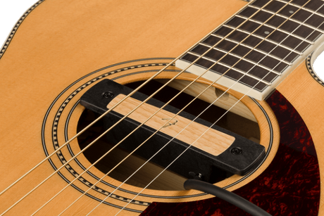The image shows a close-up of an acoustic guitar focusing on the soundhole with decorative rosette, steel or nylon strings, light-colored soundboard, dark reddish-brown pickguard, part of the fretboard with markers, and the bridge area where strings anchor. The traditional design and wood grain highlight the craftsmanship and key features that enable the guitar to produce sound.