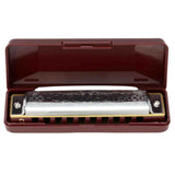 The image shows a diatonic harmonica, also known as a mouth harp or blues harp, resting in a burgundy hard plastic protective case. The harmonica features a chrome top plate with decorative engraving and a black comb with ten holes visible. The hinged case has a fitted interior to protect the instrument, which is popular for blues, folk, country, and rock music due to its portability and versatility.