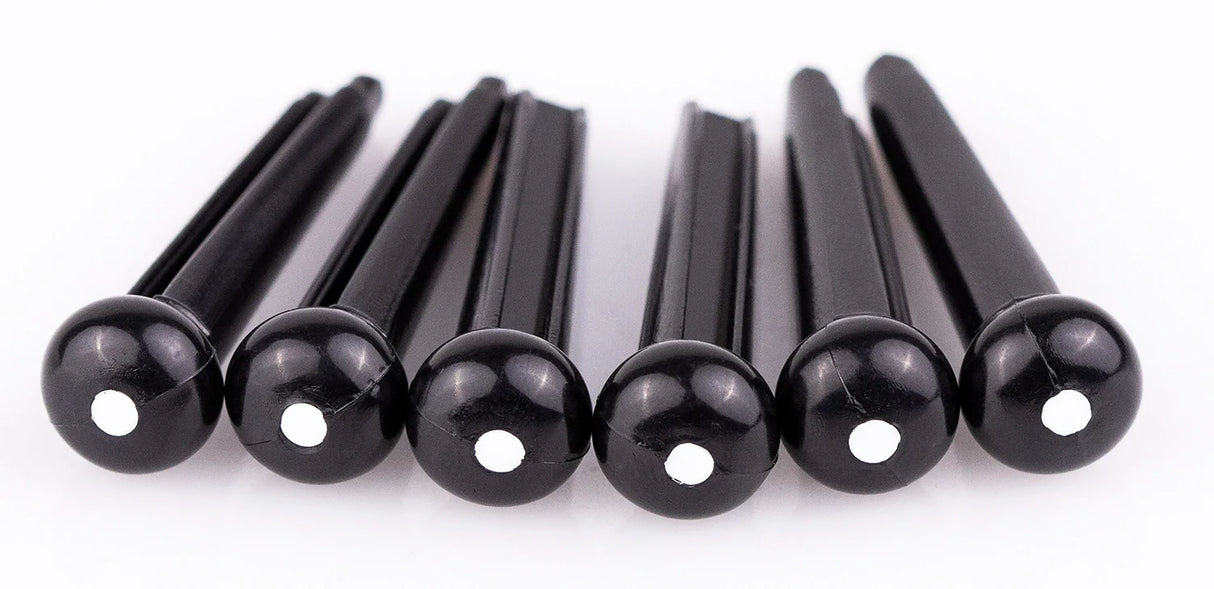 The image shows six black guitar bridge pins, likely made of plastic or ebony, each with a small white dot on the rounded end. These pins secure the strings at the bridge of an acoustic guitar by holding the string ball ends in place. Their tapered design ensures a snug fit, while the rounded tops make removal easy. They are common replacement parts for maintaining acoustic guitars.