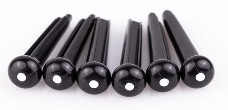 The image shows six black guitar bridge pins, likely made of plastic or ebony, each with a small white dot on the rounded end. These pins secure the strings at the bridge of an acoustic guitar by holding the string ball ends in place. Their tapered design ensures a snug fit, while the rounded tops make removal easy. They are common replacement parts for maintaining acoustic guitars.