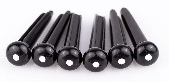 The image shows six black guitar bridge pins, likely made of plastic or ebony, each with a small white dot on the rounded end. These pins secure the strings at the bridge of an acoustic guitar by holding the string ball ends in place. Their tapered design ensures a snug fit, while the rounded tops make removal easy. They are common replacement parts for maintaining acoustic guitars.