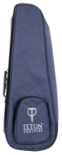 The image shows a Teton Music ukulele gig bag made of blue denim-textured fabric with a zippered edge and a small front pocket featuring the "TETON UKULELES" logo in white. Designed for concert or tenor ukuleles, this padded, soft-sided case offers lightweight protection during transport. It includes carrying handles and likely shoulder straps, making it a practical accessory for musicians on the go.