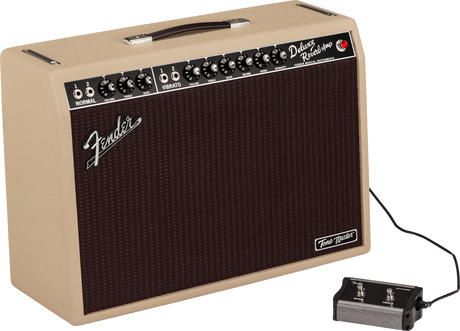 The image shows a Fender Deluxe Reverb-Amp guitar amplifier with a blonde/cream tolex exterior and dark brown grille. The control panel features multiple knobs for volume, treble, bass, reverb, and vibrato, with two input jacks labeled "NORMAL." A black footswitch is connected, and a black leather handle is on top. The "Tone Master" badge is visible on the lower right. This classic tube combo amp is known for its warm tone and built-in spring reverb.